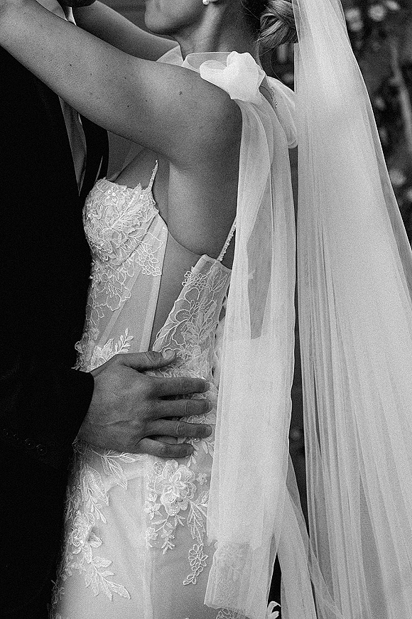 Wedding kiss as bride in a lace gown and veil embraces groom in dark suit, pearl earrings visible with blurred guests behind