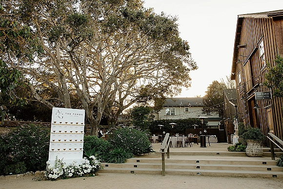 Wedding seating chart escort card display with white florals on steps under string lights in a barn courtyard with patio heaters and bistro tables