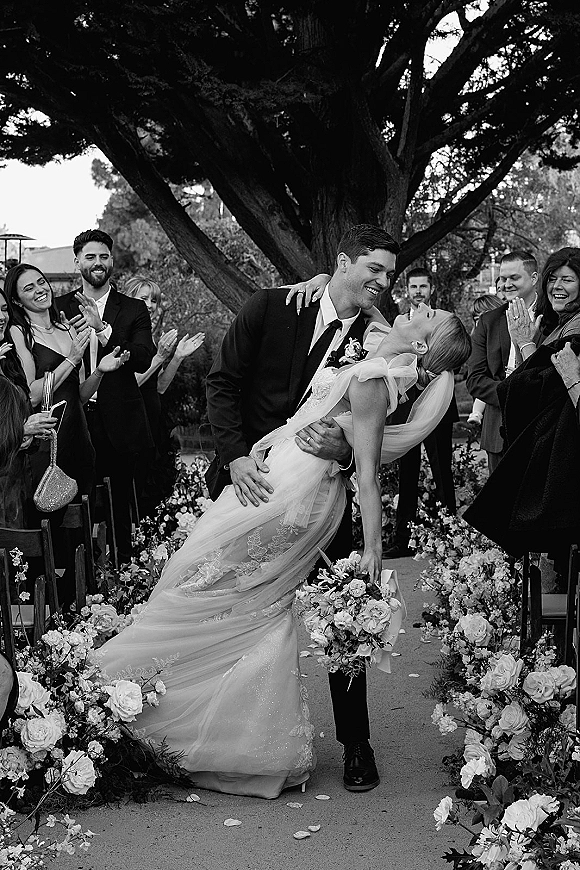 Wedding recessional just married exit with groom dipping the bride for a kiss, bouquet and veil flowing as guests cheer under a large tree