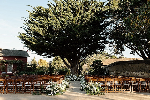 Outdoor ceremony setup with garden wedding ceremony seating, wooden chairs lining an aisle with white flowers and greenery under a large tree in a courtyard