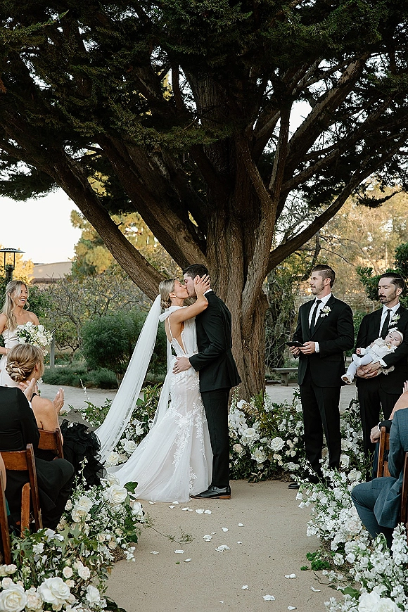 Ceremony kiss as bride in lace gown and long veil kisses groom in black suit beneath a large tree, guests watching by petal-lined aisle