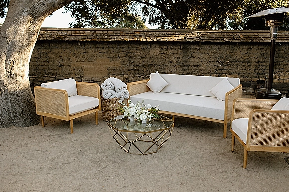 Wedding lounge seating with white cushions, rolled blankets, and a glass coffee table floral centerpiece on a patio by a stone wall