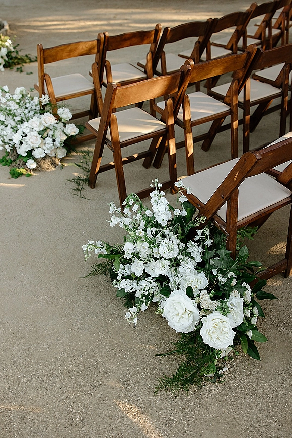 Ceremony aisle decor with wedding aisle florals lining a concrete outdoor walkway beside wood folding chairs with white cushions and greenery