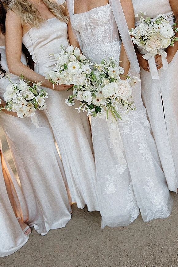 Bridesmaid bouquets of white florals with blush roses, ranunculus, anemones and greenery, held beside satin dresses on outdoor ground