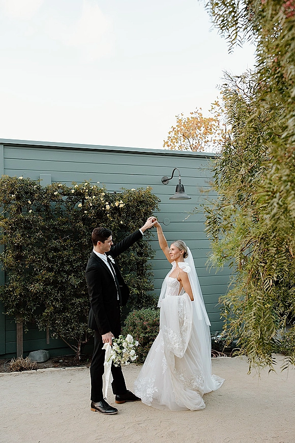 Couple portrait of bride and groom twirl as he spins her, veil and bouquet flowing beside climbing roses on garden wall siding outdoors