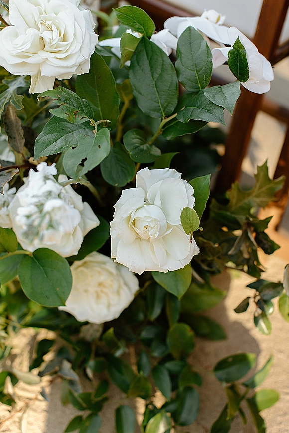 Wedding florals of white wedding flowers with white roses, orchids, and greenery draped over a wooden chair in outdoor light on sand