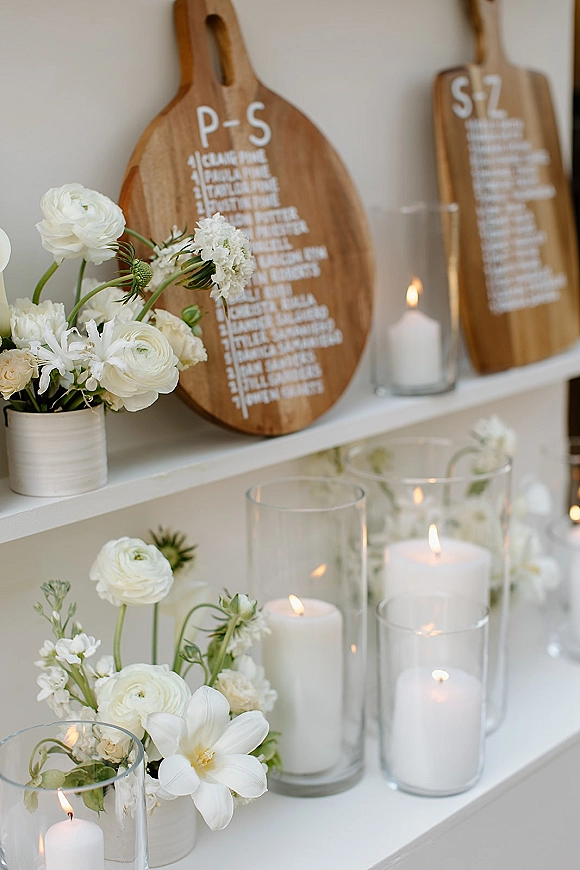 Escort card display seating chart alternative on white shelves with wood paddle boards, hand-lettered cards, white flowers, and pillar candles in glass