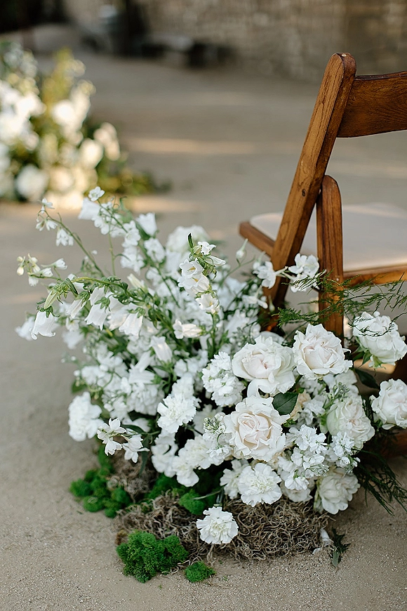 Ceremony aisle flowers in white blooms and greenery with moss at the base beside a wooden chair on a stone aisle near a brick wall