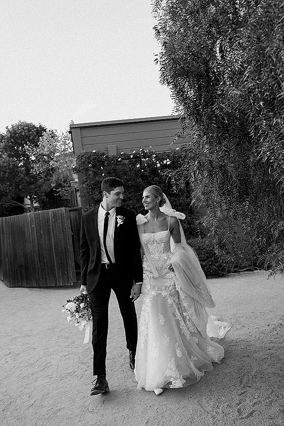 Couple portrait of bride and groom walking hand in hand on a gravel path, bride looking at groom, veil flowing, bouquet in hand