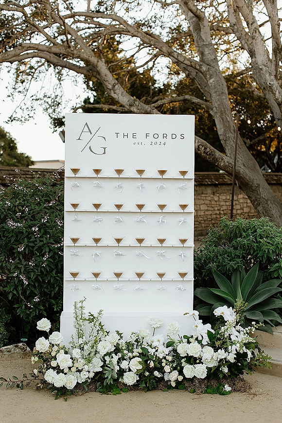 Wedding escort display with a martini escort wall of cocktail drinks on a white monogram board, with rose florals in a garden setting