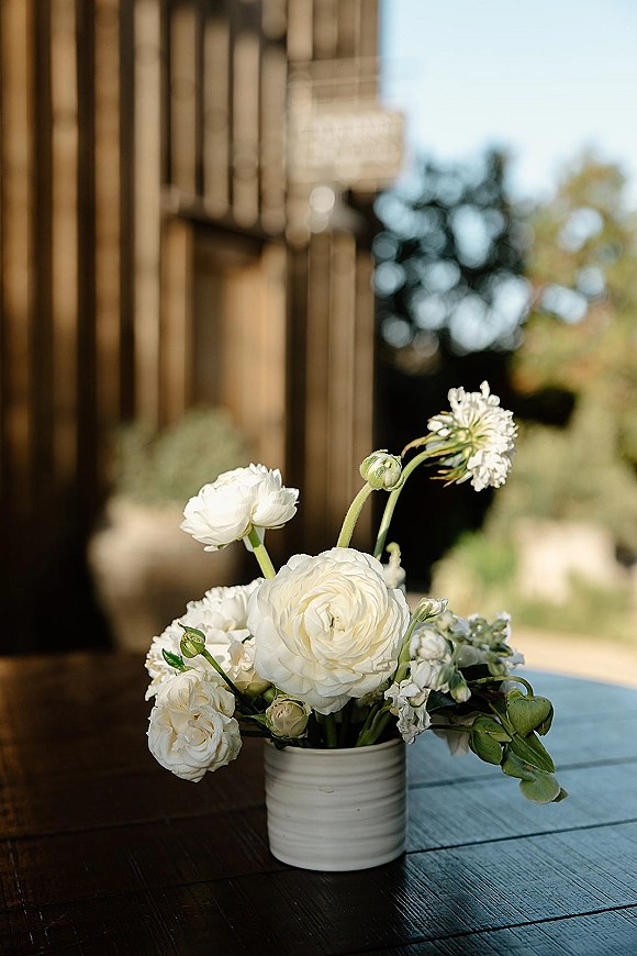 Wedding centerpiece with white flower centerpiece in a ceramic vase on a wood table, set outside near a wooden building and greenery