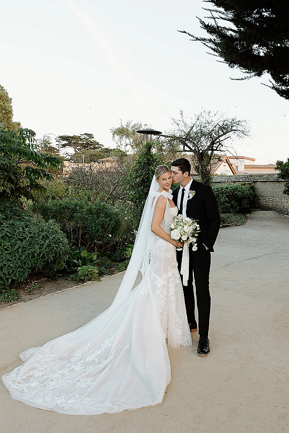 Couple portrait of groom kissing bride’s forehead as she holds a white bouquet, long veil and lace train on a garden path