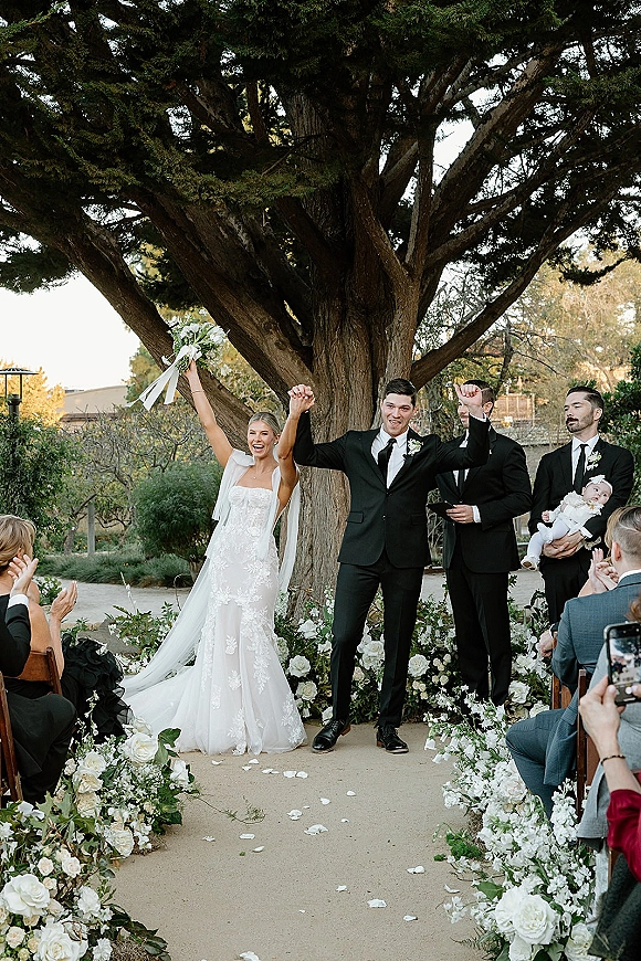 Wedding recessional as bride and groom walk up the aisle with raised hands, bride lifting bouquet under a large tree in a garden courtyard