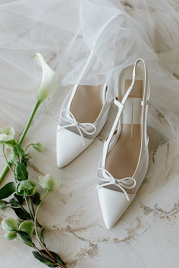 Bridal shoes with bow details, white wedding heels with ankle straps and mesh panels on marble beside a tulle veil and calla lily
