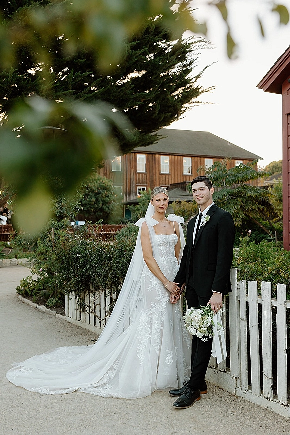 Couple portrait of bride and groom holding hands, her long veil and bouquet beside a white picket fence on a garden path