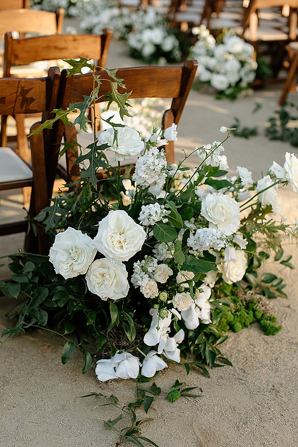 Ceremony aisle decor with aisle floral arrangement of white roses and greenery lining a concrete walkway beside wooden chairs outdoors