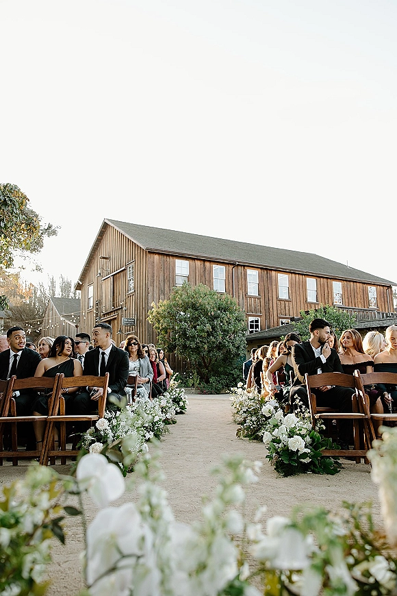 Ceremony aisle decor with white floral arrangements and greenery garlands lining wooden folding chairs beneath string lights in a rustic barn courtyard