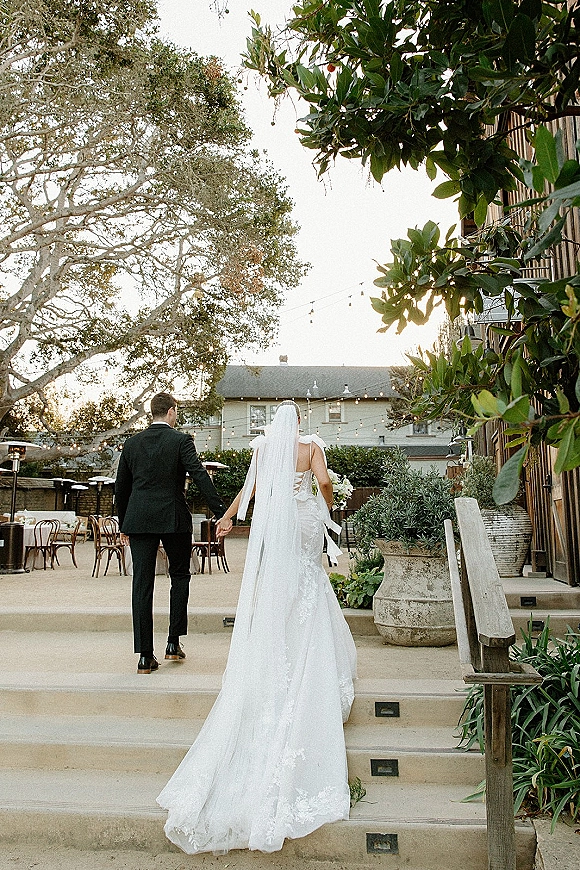 Couple portrait of bride and groom walking away holding hands, her long veil trailing, under string lights in a rustic courtyard