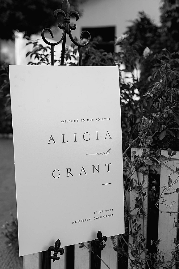 Wedding welcome sign with calligraphy lettering on a wrought iron stand along an outdoor walkway with greenery and a fence backdrop