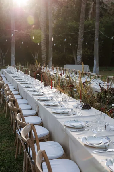 Reception tablescape with a long banquet table, white linens, taper candles and greenery runner, set under string lights near palm trees