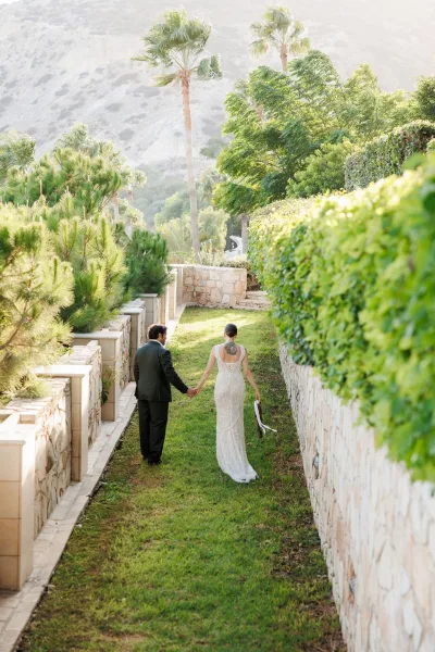 Couple portrait of newlyweds walking away holding hands, bride lifting a long train with tattoo visible on a garden path by stone wall and palms