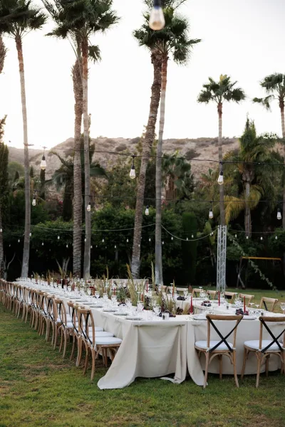 Reception tablescape at an outdoor wedding reception with a long banquet table, white linens, taper candles, greenery, and bistro lights under palm trees