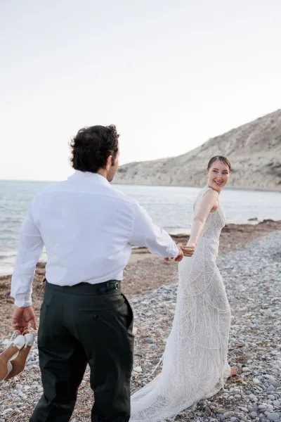 Couple portrait of bride and groom holding hands, walking on a rocky beach as she looks back, ocean cliffs behind them
