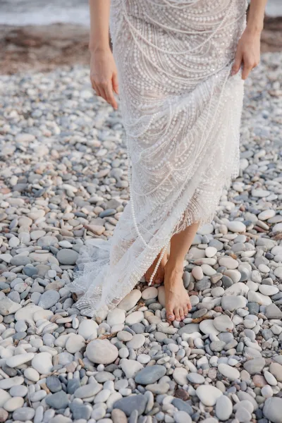 Wedding dress detail with beaded wedding dress lace and sequins, sheer tulle hem brushing shoreline stones as the bride stands barefoot on a pebble beach