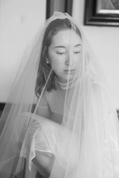 Bridal portrait in black and white of a bride looking down, wedding veil draped over her face, lit by window light near wall paneling