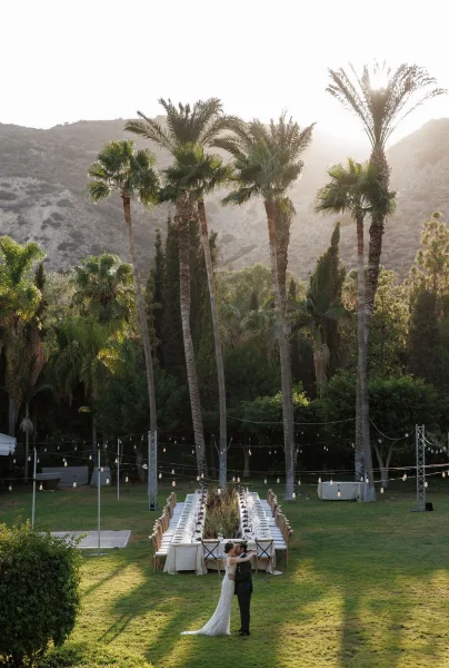 Reception tablescape with a long banquet table wedding setup, white linens, cross-back chairs, floral centerpieces and string lights at sunset.