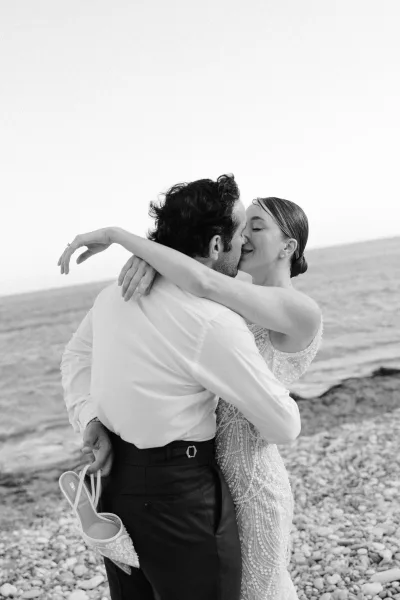 Wedding kiss portrait of a beach wedding couple embracing on a rocky shoreline, bride in a beaded dress with stud earrings by the ocean horizon