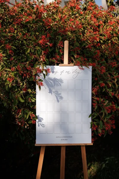 Wedding seating chart on a white board atop a wooden easel with calligraphy text, set against a flowering hedge in an outdoor garden