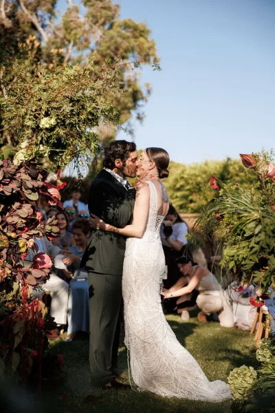 Wedding kiss under a floral arch, bride in a beaded wedding dress and groom in a black suit as guests cheer on the garden lawn