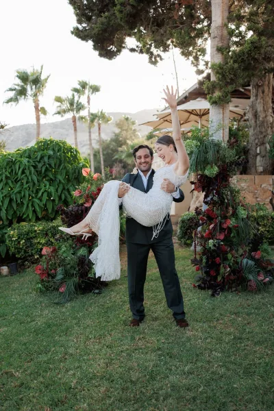 Couple portrait of groom lifting bride in a wedding lift pose beneath a modern anthurium floral arch on a garden lawn with mountains behind