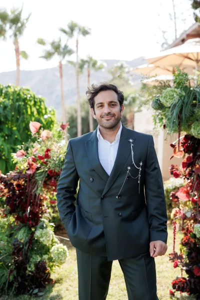 Groom portrait in a double breasted suit with boutonniere chain, standing before garden florals with palm trees and mountains backdrop