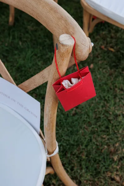 Wedding favor bag in bright red with drawstring handles hanging from a wooden ceremony chair beside a printed program on a grass lawn