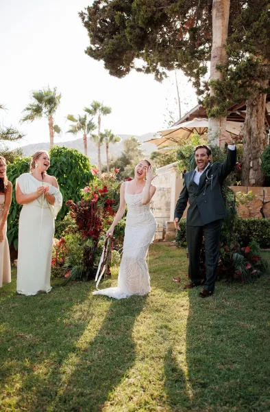 Wedding recessional as bride and groom exit laughing, bride in beaded dress with bouquet ribbon streamers, guests cheering on a sunny garden lawn with palm trees