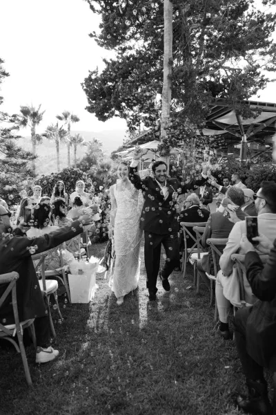 Wedding recessional as bride and groom walk the aisle through a bubble wedding exit, guests cheering among chairs and florals outdoors.