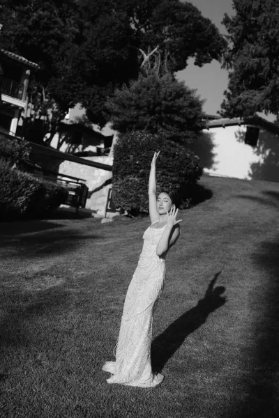 Bridal portrait in black and white of a full-length bride in a beaded spaghetti-strap gown showing her ring on a lawn by trees and hillside house