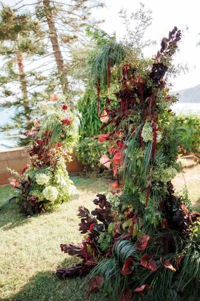 Wedding ceremony arch with asymmetrical wedding arch florals, hydrangeas and burgundy blooms on a lawn by a lake and mountains in back