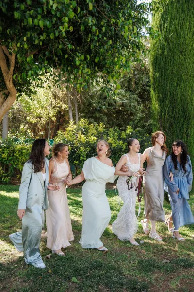 Bridesmaids group photo with mismatched dresses and colorful bouquets, holding hands and laughing on a sunlit garden lawn