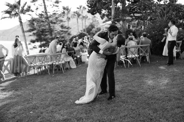 Wedding kiss portrait as groom dips the bride in her gown and veil while guests watch on a lawn with palm trees and ocean view
