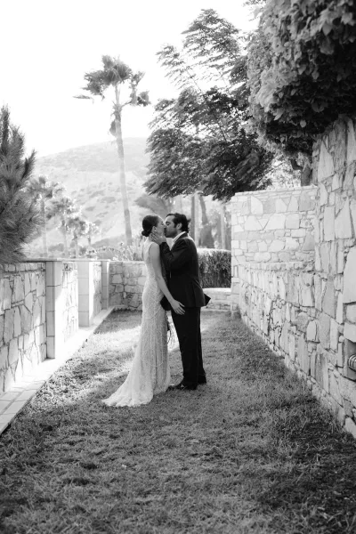 Wedding kiss as bride and groom embrace, veil and earrings visible beside stone walls and palm trees with mountains beyond
