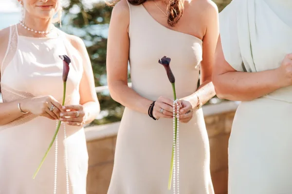 Bridesmaid dresses in mismatched neutral tones with hands holding calla lily bouquets and pearl jewelry on a terrace railing by trees