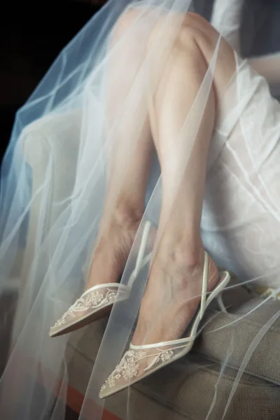 Bridal shoes with lace wedding heels detail, pointed toe slingbacks draped in tulle veil on an upholstered chair