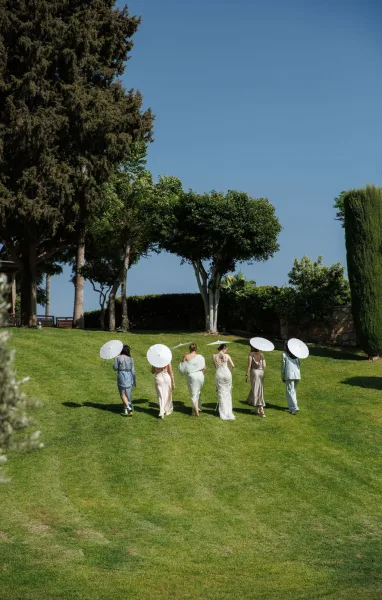 Bridal party walking across a sunny lawn, bridesmaids with parasols behind the bride in a white gown, with trees and stone wall backdrop