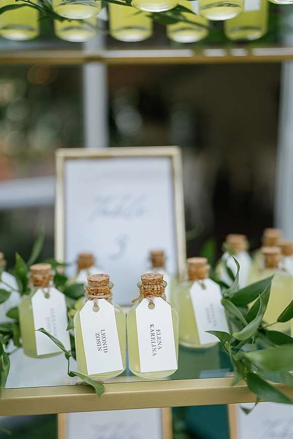 Wedding place cards on glass bottles with cork stoppers and white name tags, arranged on a shelf with greenery garland by a window