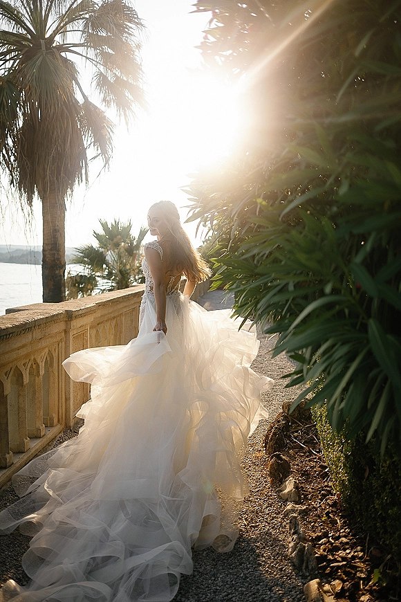 Bridal portrait of a bride looking over her shoulder in a backless wedding dress with lace bodice and tulle train on a palm-lined terrace