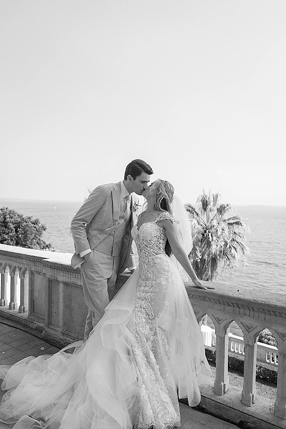 Wedding kiss portrait of bride and groom kissing on a stone balcony, her lace dress and cathedral veil flowing with ocean and palm trees behind