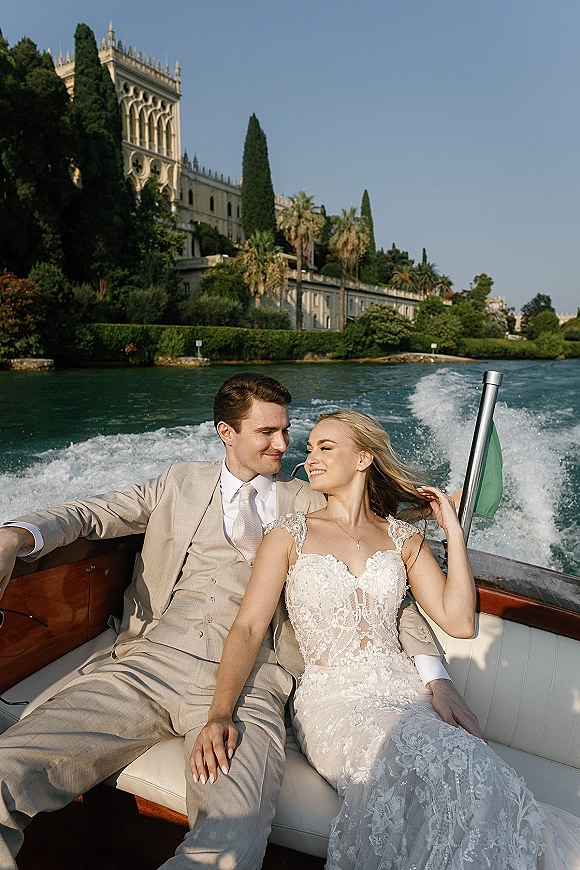 Couple portrait on a wooden motorboat, bride in a lace wedding dress and groom in a beige suit, cruising a lake past villas and palms